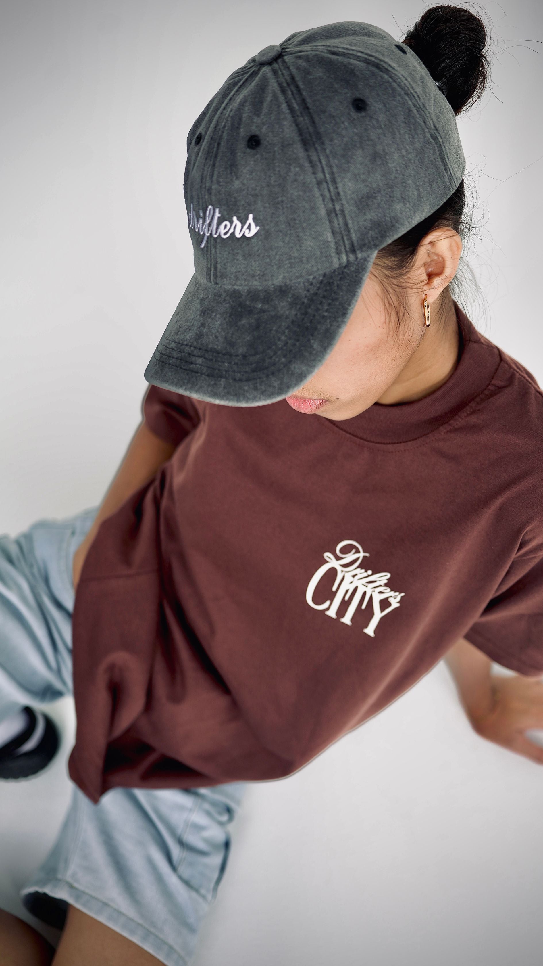Person wearing a gray cap and brown shirt with text, sitting on a white surface.